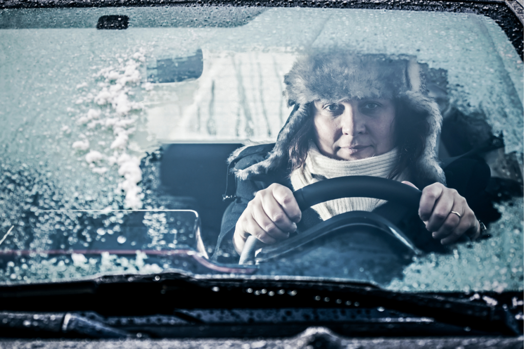 Person sitting in a car on a winter day, holding the steering wheel while looking through a partially iced-over windshield.