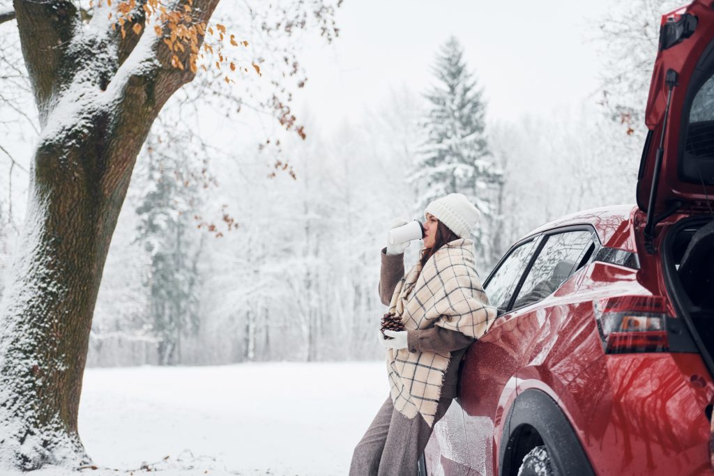 Person leaning against a red SUV in a snowy forest, wrapped in a blanket and drinking from a mug.